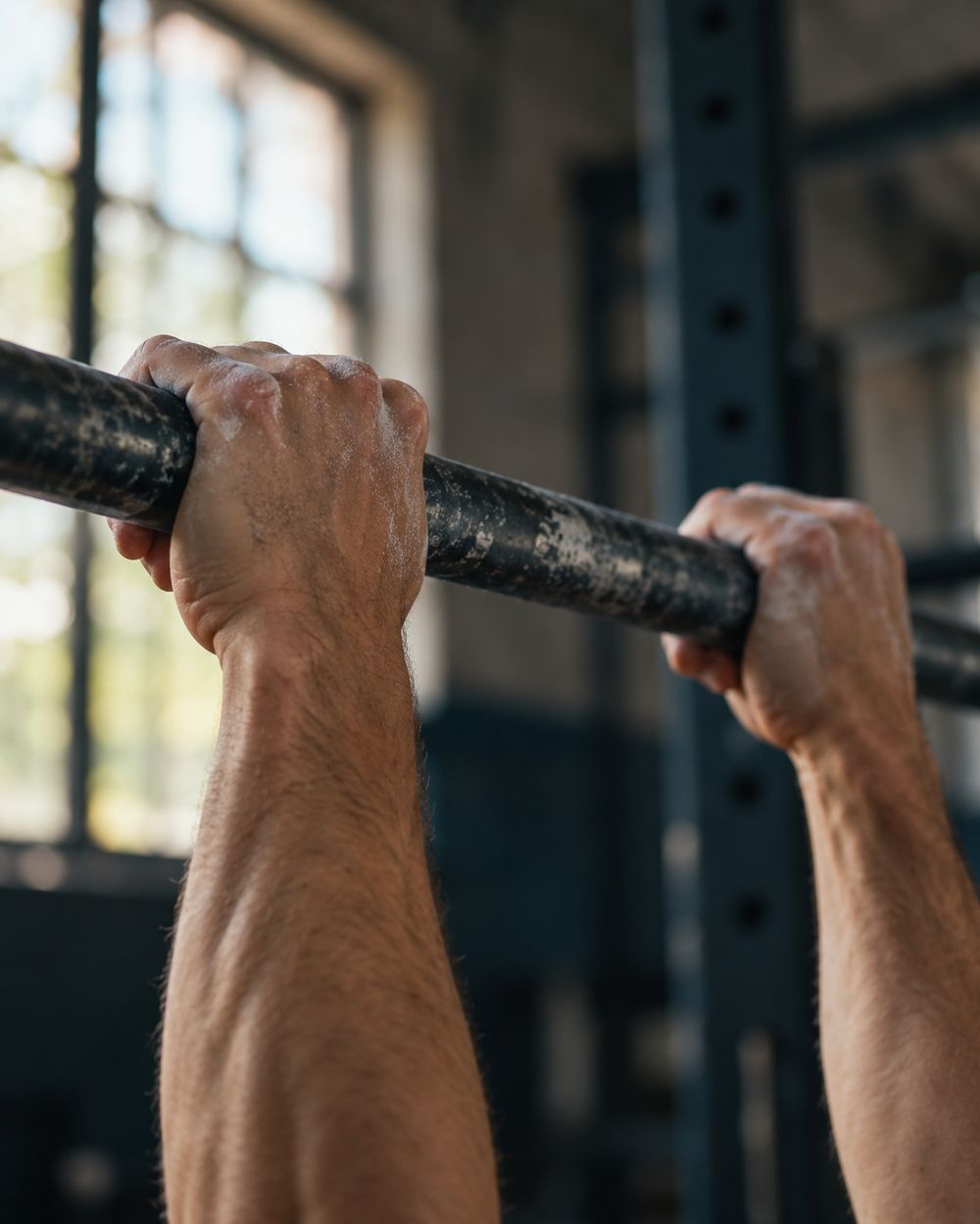 Close-up of two chalked hands gripping a steel bar, with a power rack and a window blurred behind them.